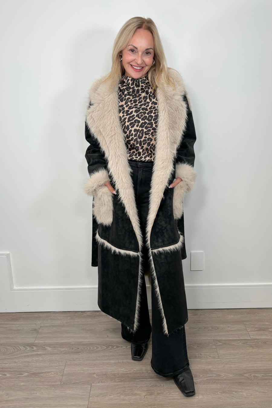 Front view of a woman wearing an open long black coat with fur detailing and hem on the collar, pockets and belt.