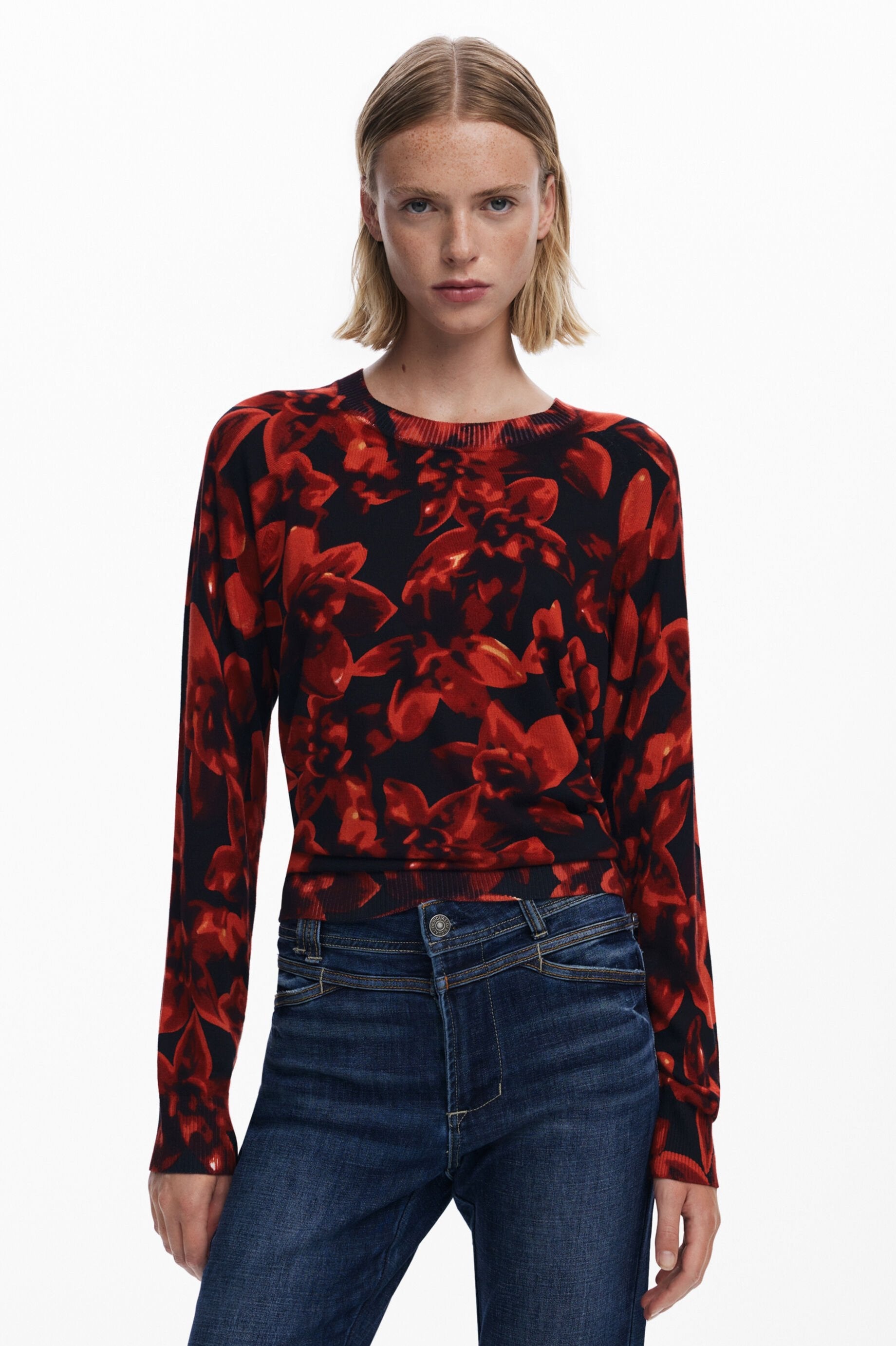 Woman wearing a red and black floral long-sleeve top and blue jeans on a white background