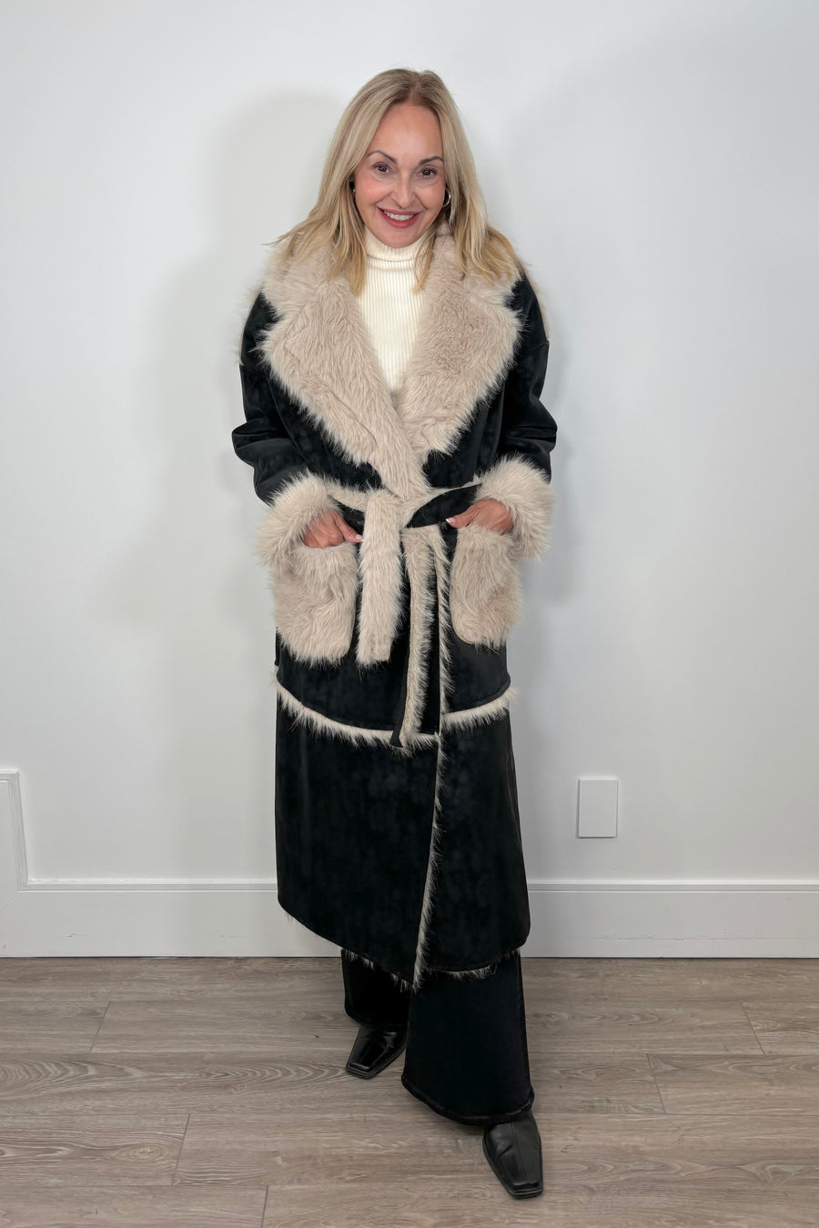 Front view of a long black coat with fur detailing and hem on the collar, pockets and belt. It is tied up