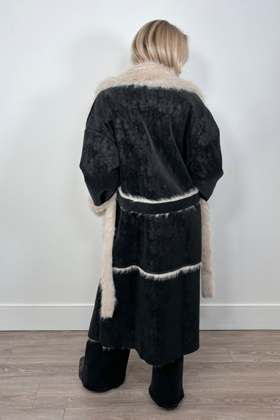 Back view of a woman wearing an open long black coat with fur detailing and hem on the collar, pockets and belt.