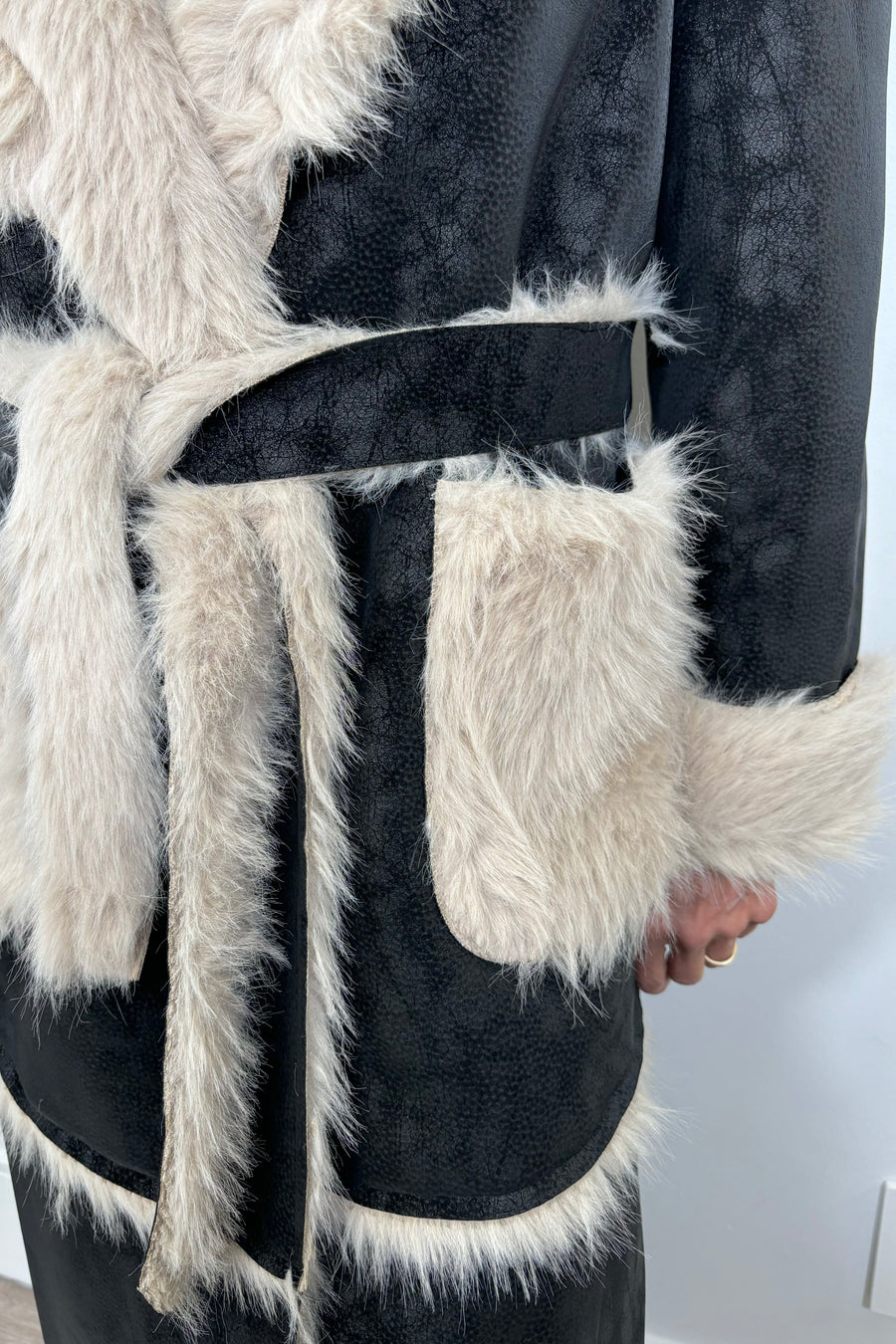 Close up view of the belt of an open long black coat with fur detailing and hem on the collar, pockets and belt.