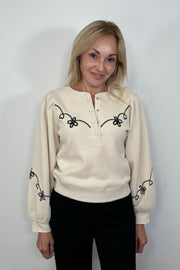 Front facing woman wearing a ecru coloured sweatshirt with a western inspired detailing in the arms and chest and Henley neck