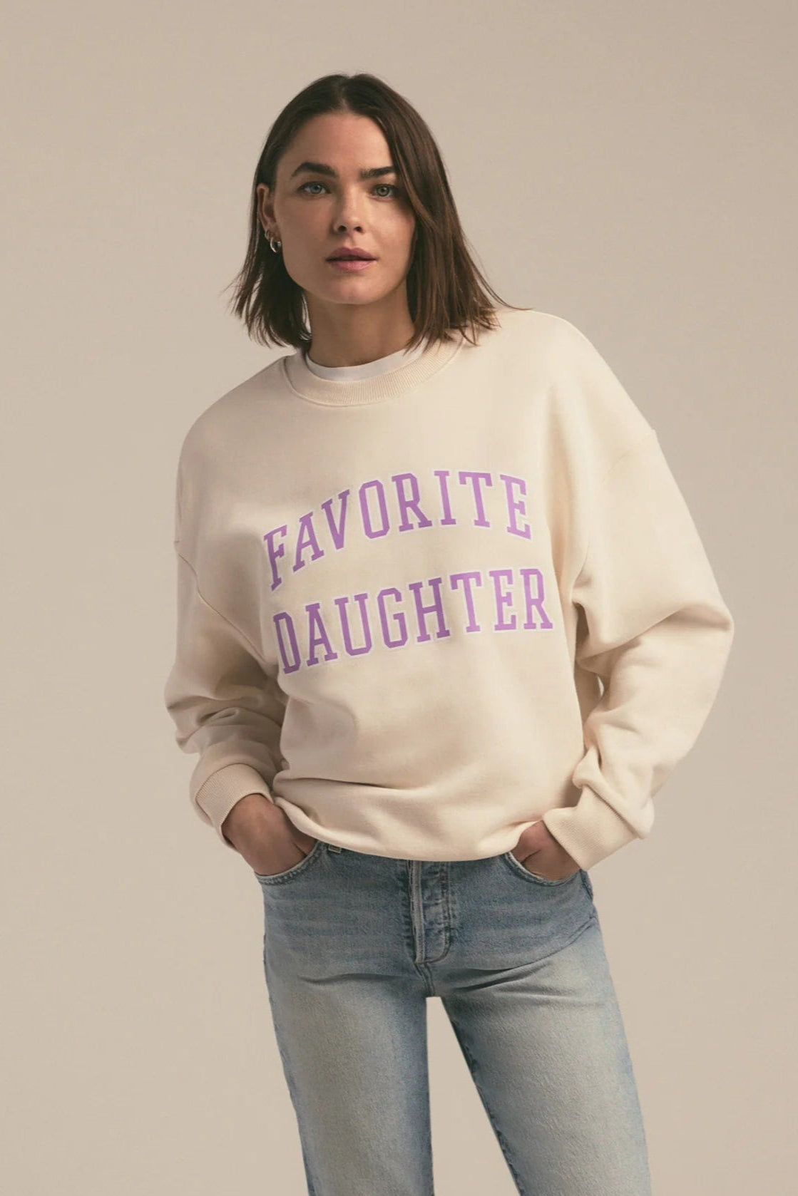 Woman wearing a beige sweatshirt with 'FAVORITE DAUGHTER' text in purple on a plain background