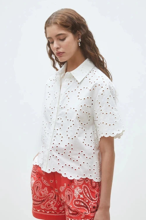 woman wearing a white short-sleeve eyelet blouse with a button-front design paired with red patterned shorts, front