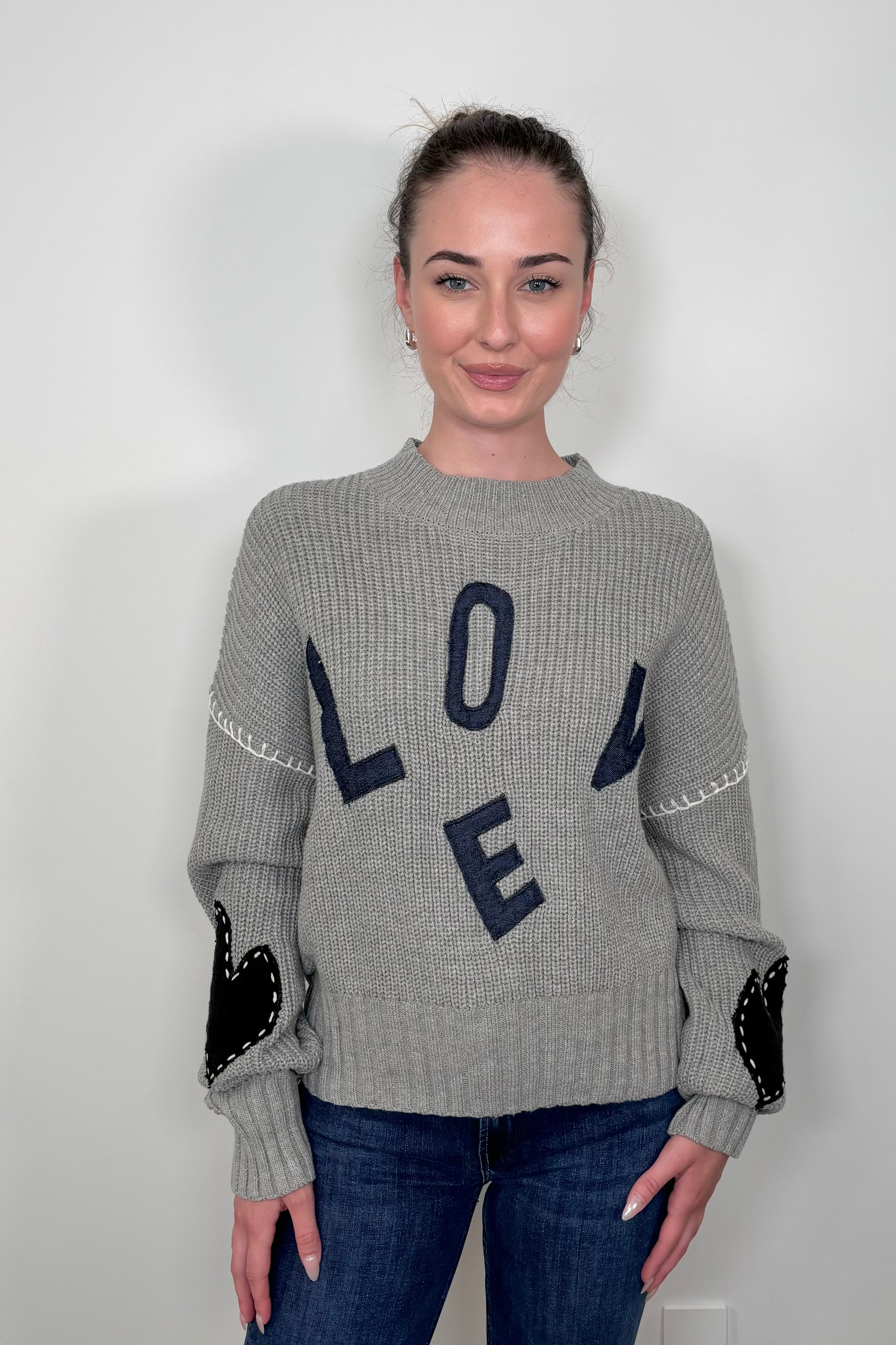 Front view of woman wearing a gray sweater with letter spelling love, hearts in the elbows with whipstitch detailing