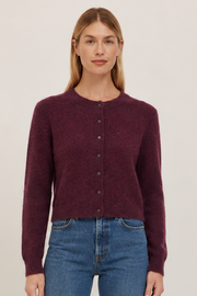 A woman stands against a plain light beige studio background wearing a dark burgundy button-front cardigan,