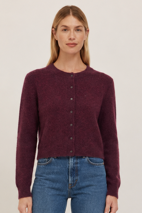A woman stands against a plain light beige studio background wearing a dark burgundy button-front cardigan,