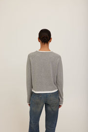 A model wearing a grey long-sleeve crewneck sweater with white ribbed trim at the collar, cuffs, and hem. 
