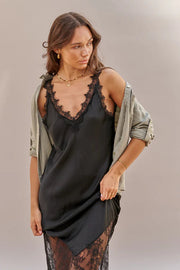 Woman wearing a black slip dress with lace details and an olive green cardigan on a neutral background