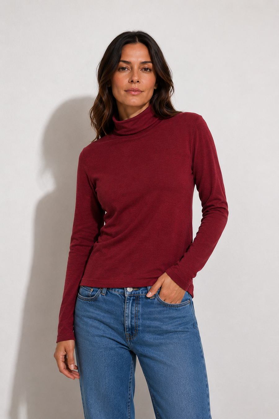 A woman modeling a long-sleeve ribbed turtleneck top in deep burgundy, paired with blue jeans, against a plain light gray studio background.