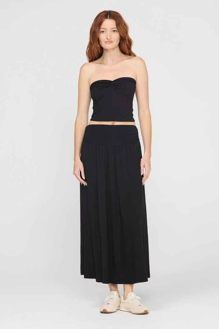 A woman modeling a black strapless bandeau-style crop top and matching black midi skirt against a light gray studio background.