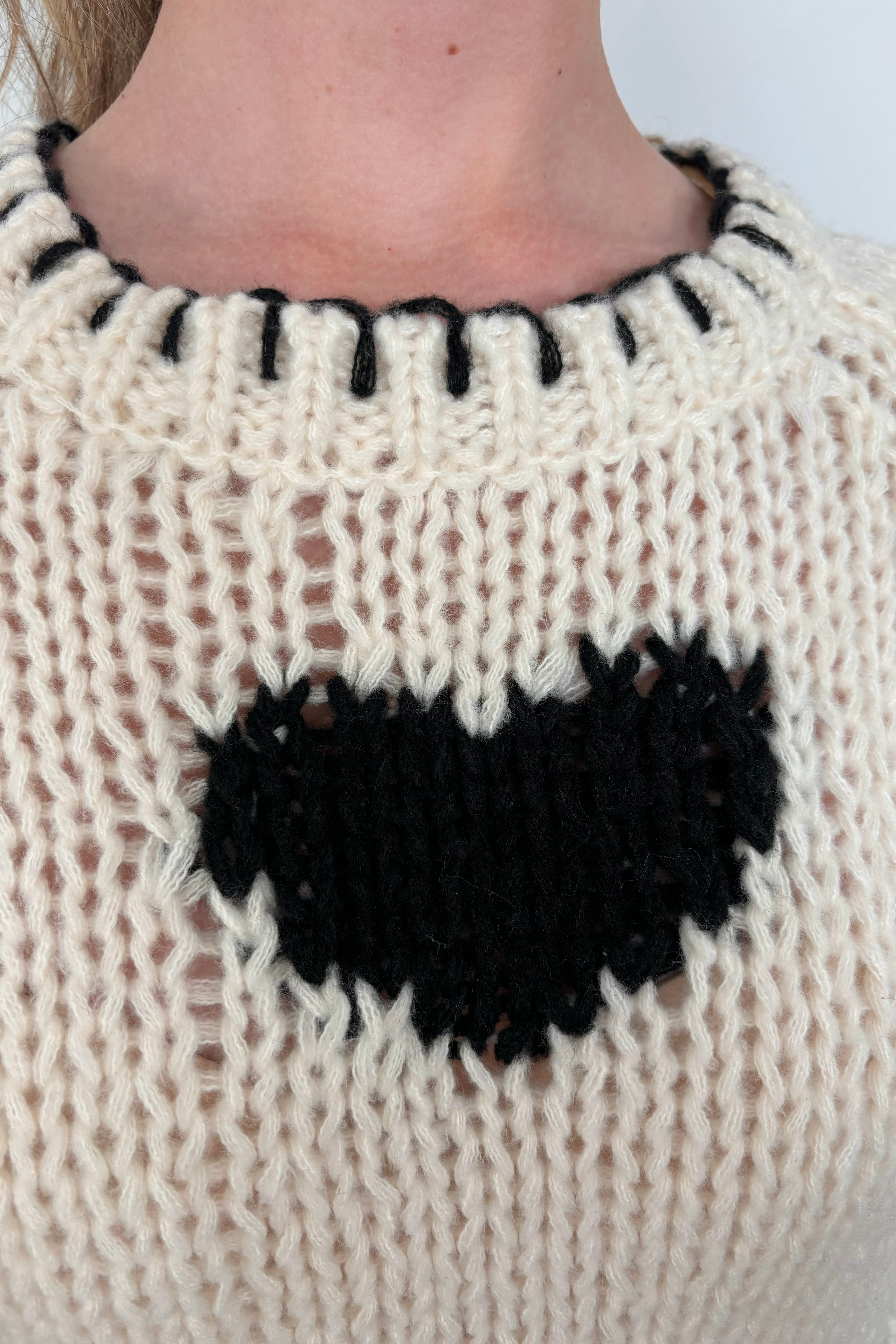 Close-up of a person wearing a cream-colored sweater with a black heart design and whip-stitch detailing on the collar