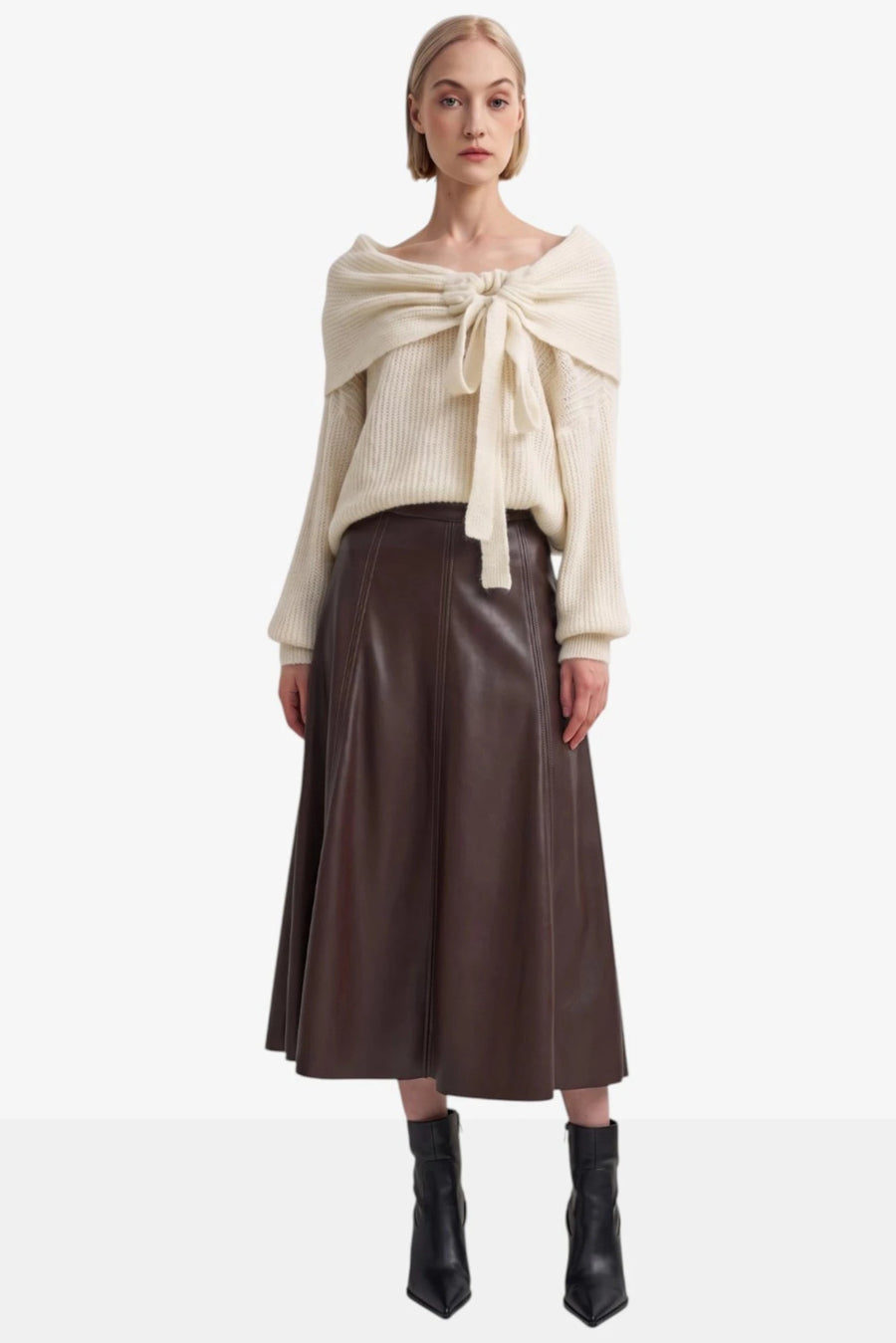 A model stands against a light gray studio background wearing a cream ribbed knit sweater with a large front bow, a dark brown midi skirt, and black ankle boots.