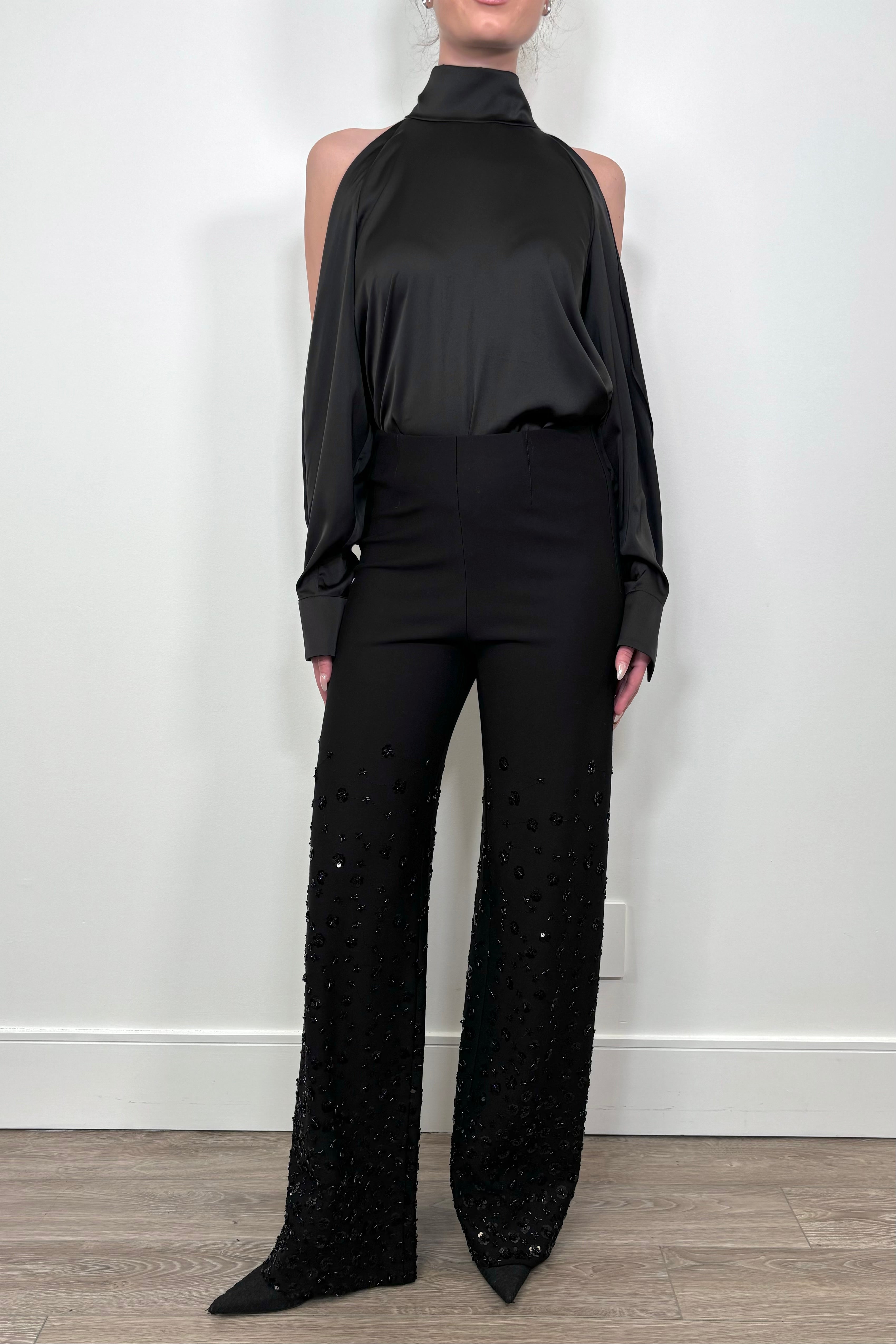 Person wearing a black outfit with a high-neck top and wide-leg pants against a white wall.