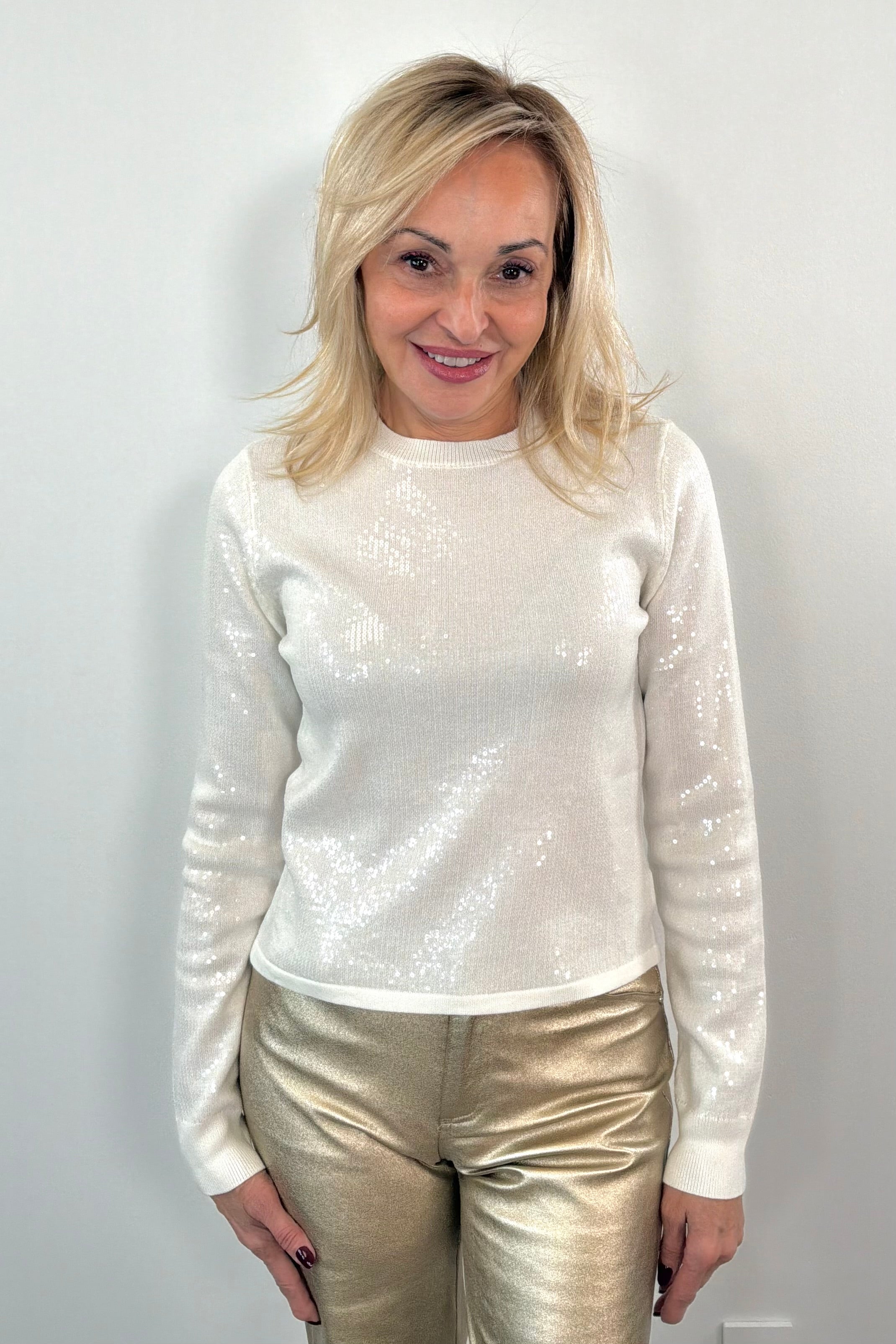 Woman wearing a white sequin top and gold pants against a plain background