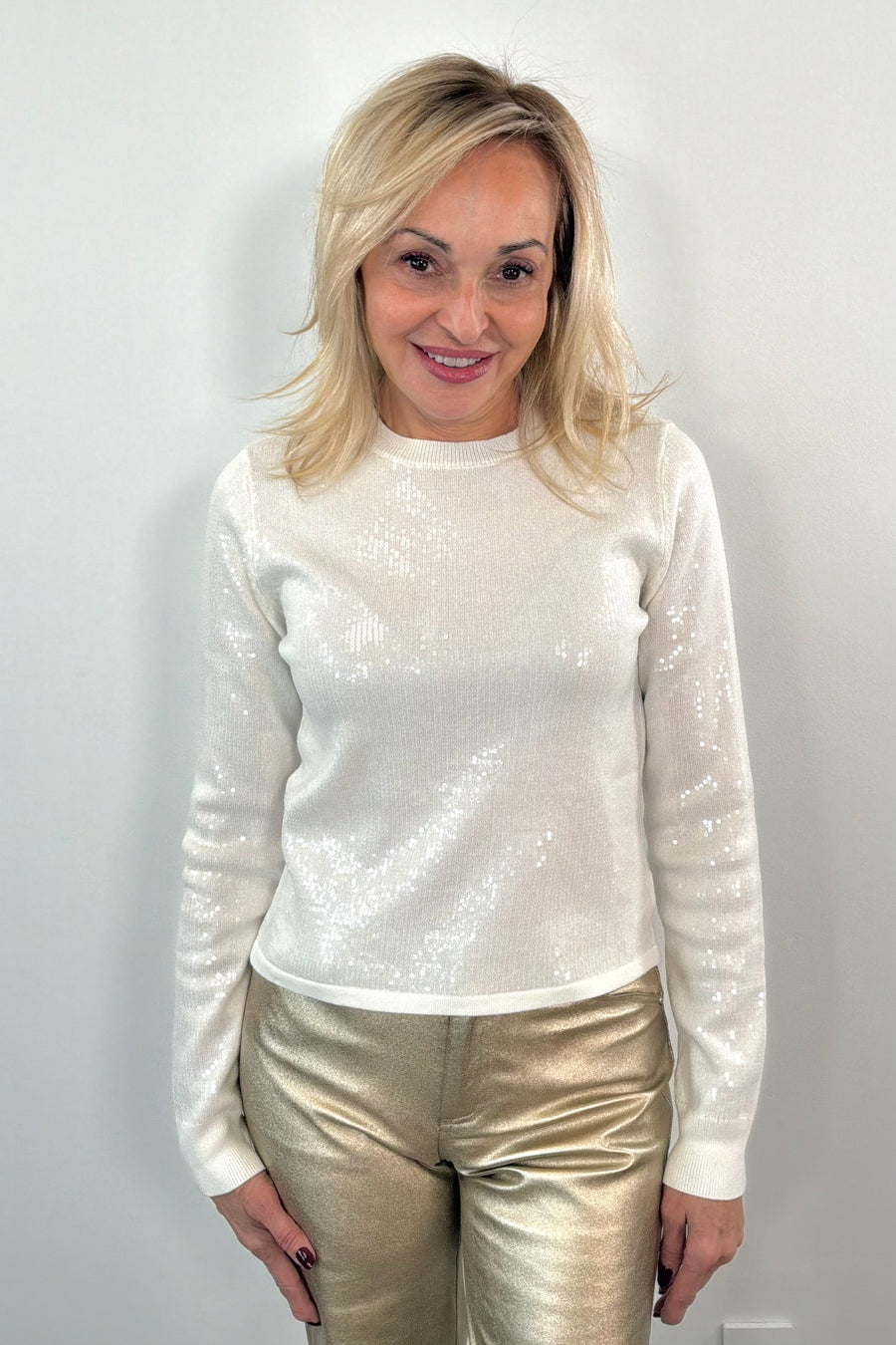 Woman wearing a white sequin top and gold pants against a plain background