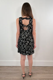 Woman wearing a black floral dress with cut-out back design against a white wall.