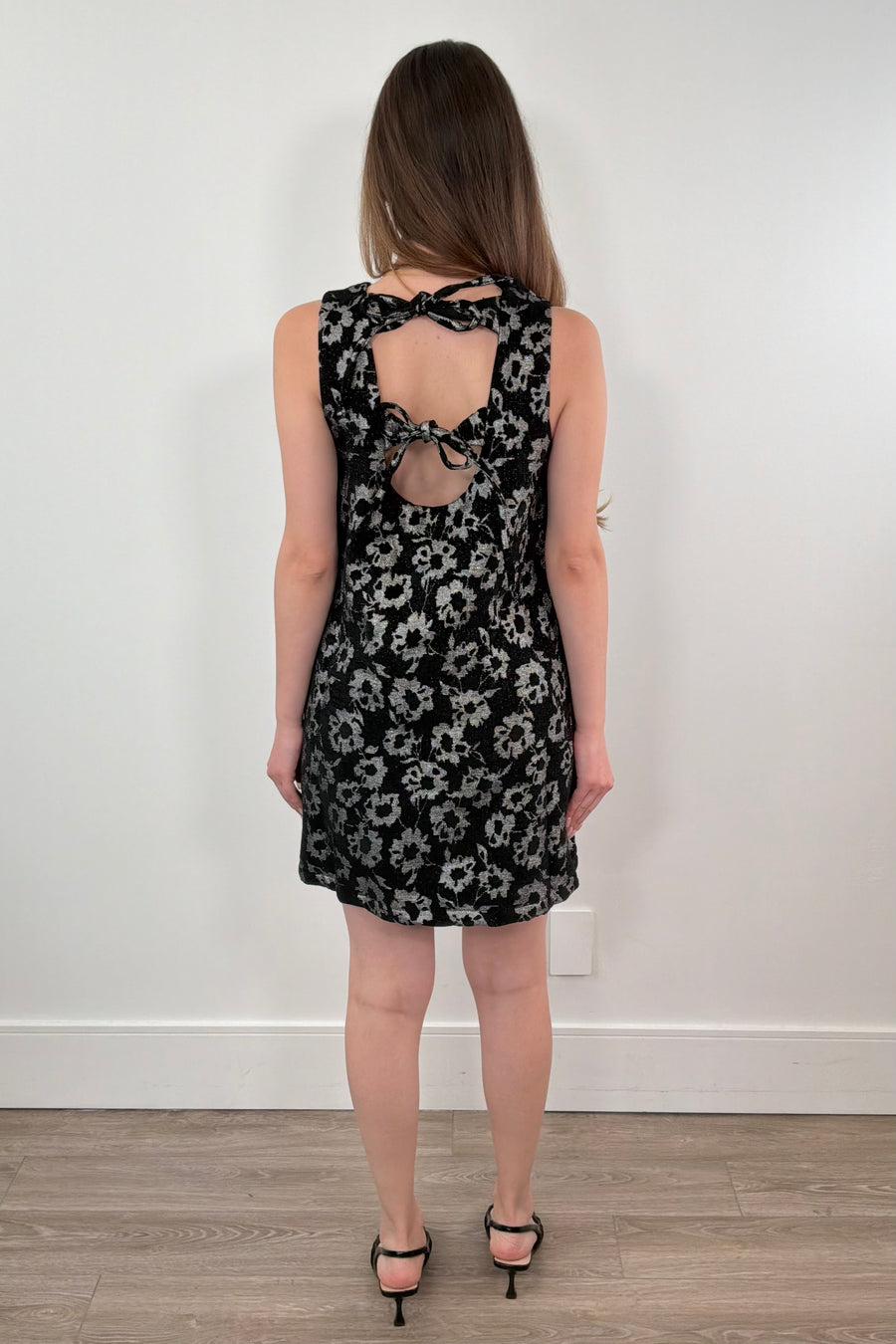 Woman wearing a black floral dress with cut-out back design against a white wall.