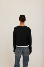A model wearing a black long-sleeve crewneck sweater with white ribbed trim at the collar, cuffs, and hem. 