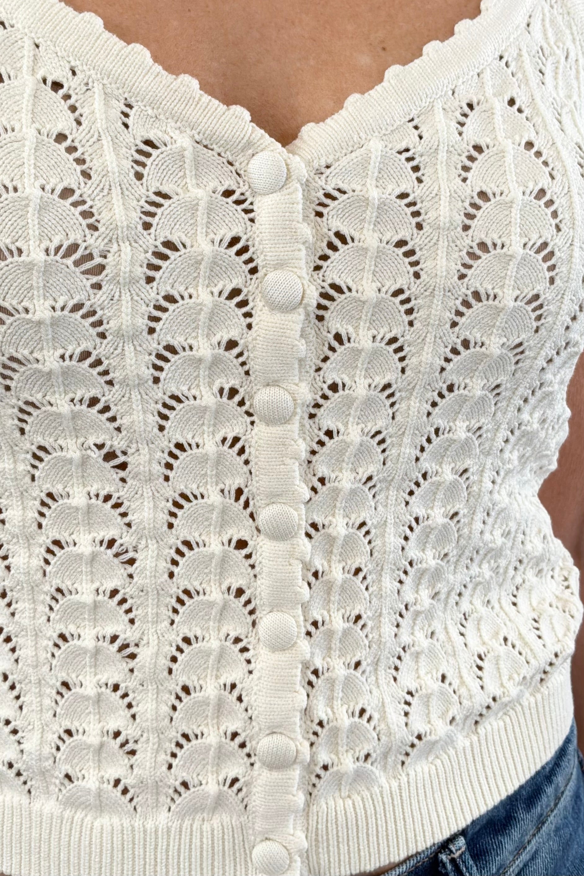 Close up of Woman wearing a cream knit camisole with buttons on the front, scalloped trim and open stitch detailing