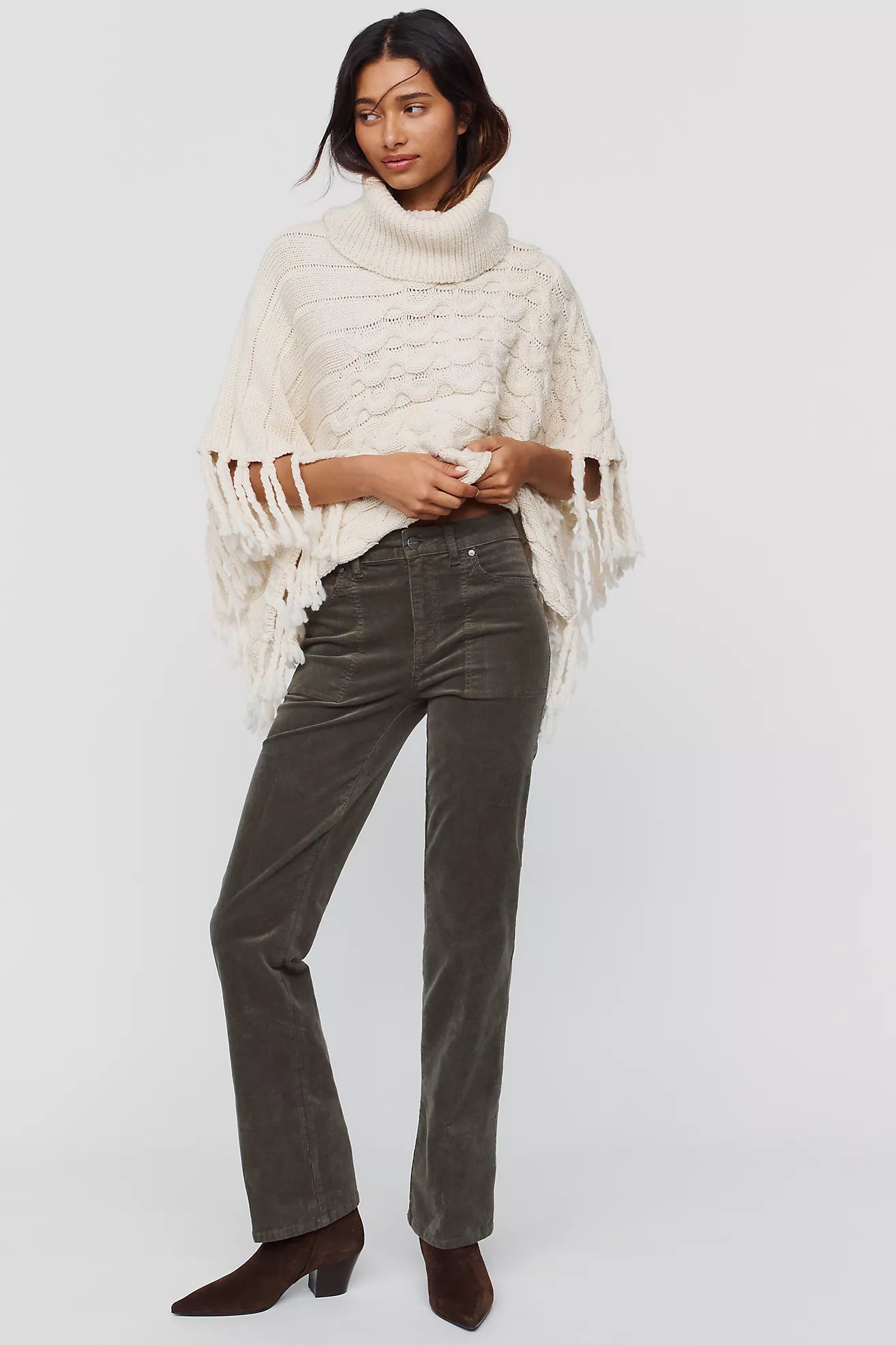 A model wearing dark olive green corduroy straight-leg jeans, styled with a cream fringed poncho and brown suede ankle boots, against a light gray studio background.