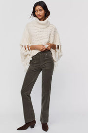 A model wearing dark olive green corduroy straight-leg jeans, styled with a cream fringed poncho and brown suede ankle boots, against a light gray studio background.