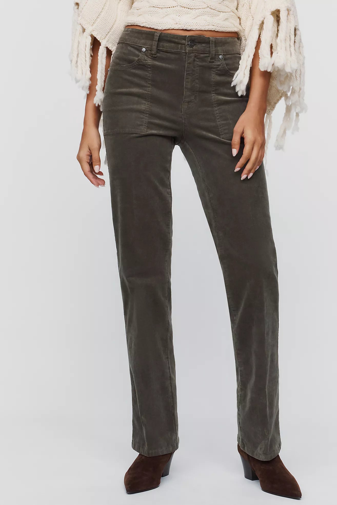A model wearing dark olive green corduroy straight-leg jeans, styled with a cream fringed poncho and brown suede ankle boots, against a light gray studio background.