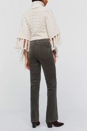 A model shown from the back wearing a cream cable-knit turtleneck poncho with fringe and dark olive green corduroy pants, against a light gray studio background.
