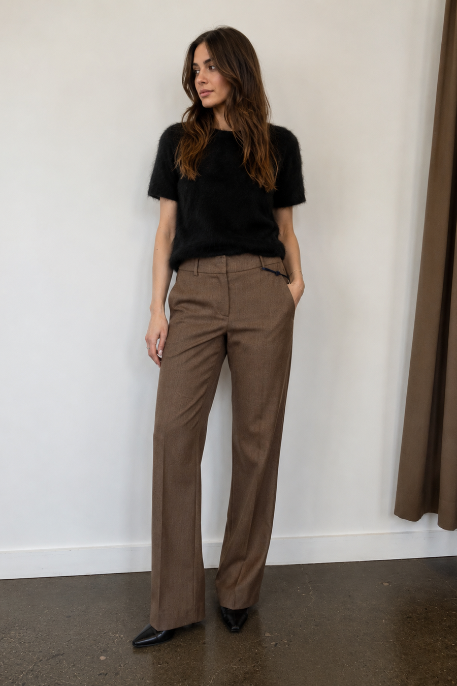 A woman stands indoors wearing a black short-sleeve top and brown straight-leg trousers