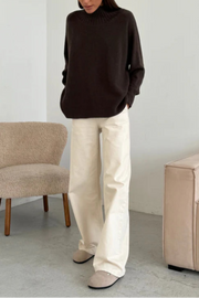 Person wearing a dark brown sweater and white pants in a minimalistic room.
