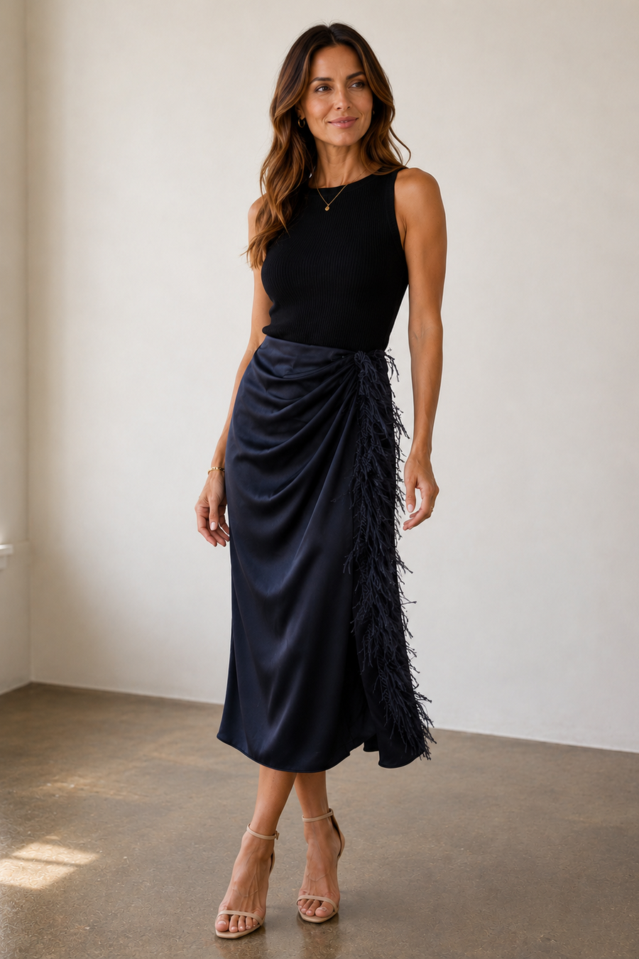 A woman modeling a sleeveless black top and a dark navy midi skirt with feathered detailing on the side, standing in a minimalist indoor room with light walls and a concrete floor.