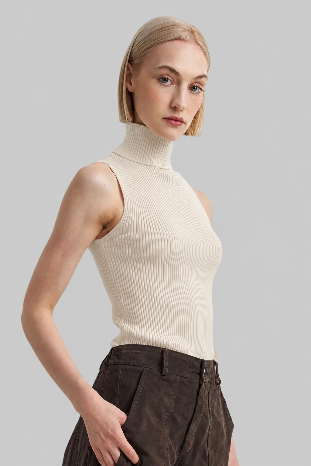 A blonde model wearing a ribbed sleeveless mock neck top in cream and dark brown trousers, photographed against a solid light gray background.