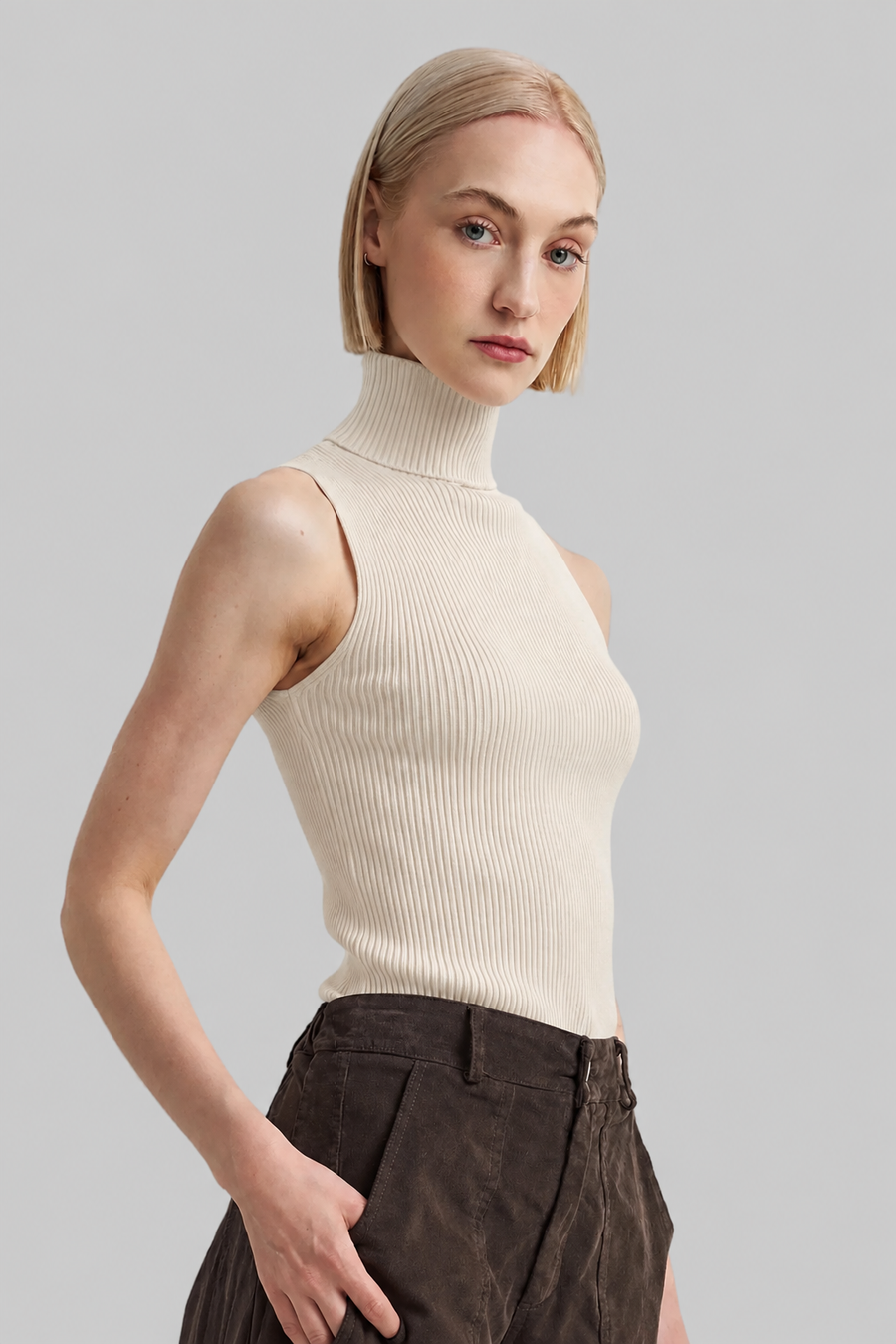 A blonde model wearing a ribbed sleeveless mock neck top in cream and dark brown trousers, photographed against a solid light gray background.
