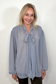 Front view of woman wearing a thin blue blouse with a bow untucked 