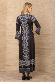 Woman wearing a long black dress with white and blue floral patterns against a wooden paneled wall.