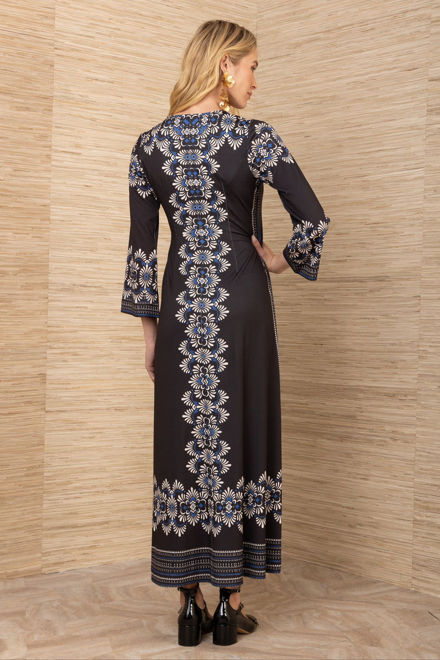 Woman wearing a long black dress with white and blue floral patterns against a wooden paneled wall.