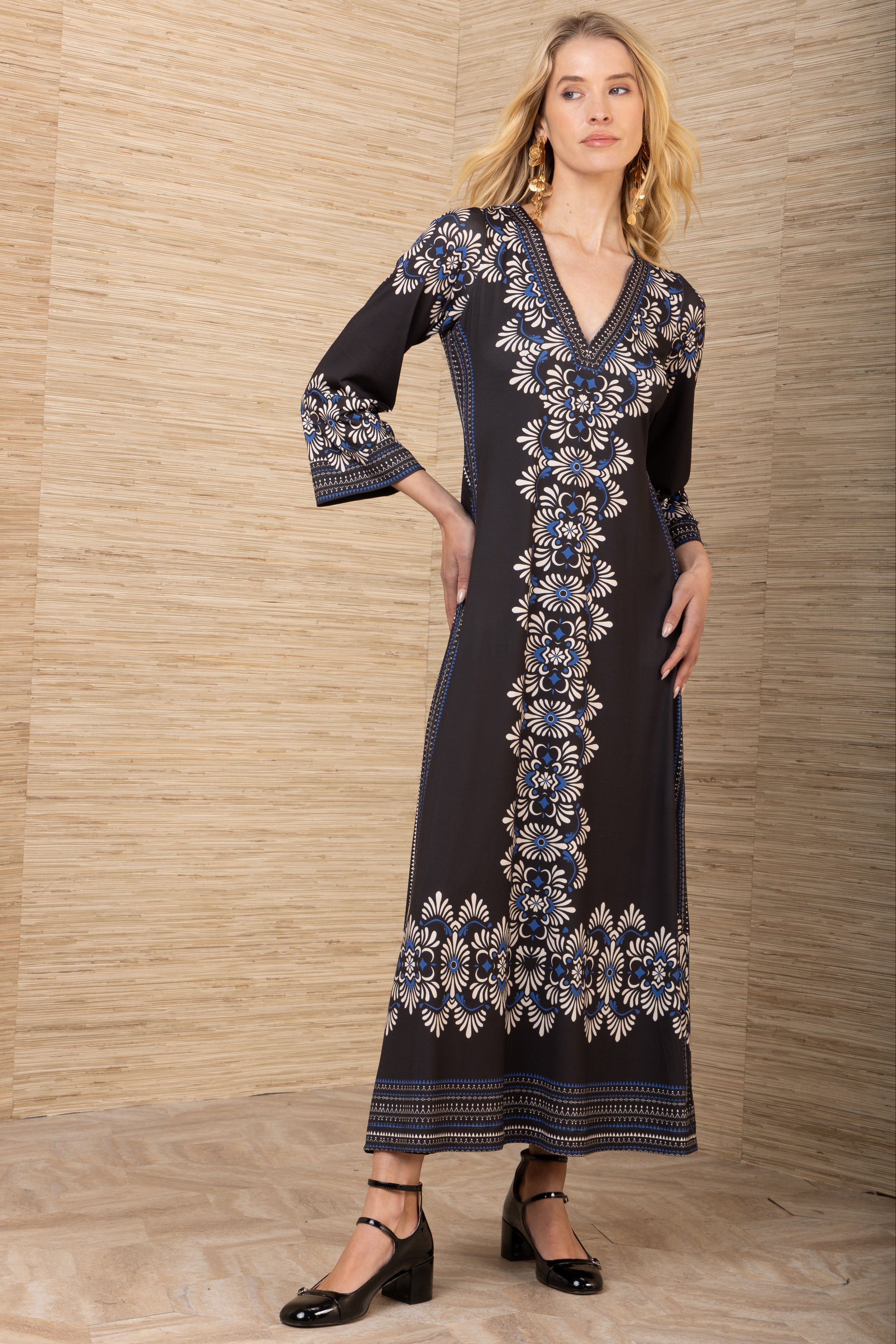 Woman wearing a black dress with blue floral embroidery against a wooden paneled wall.