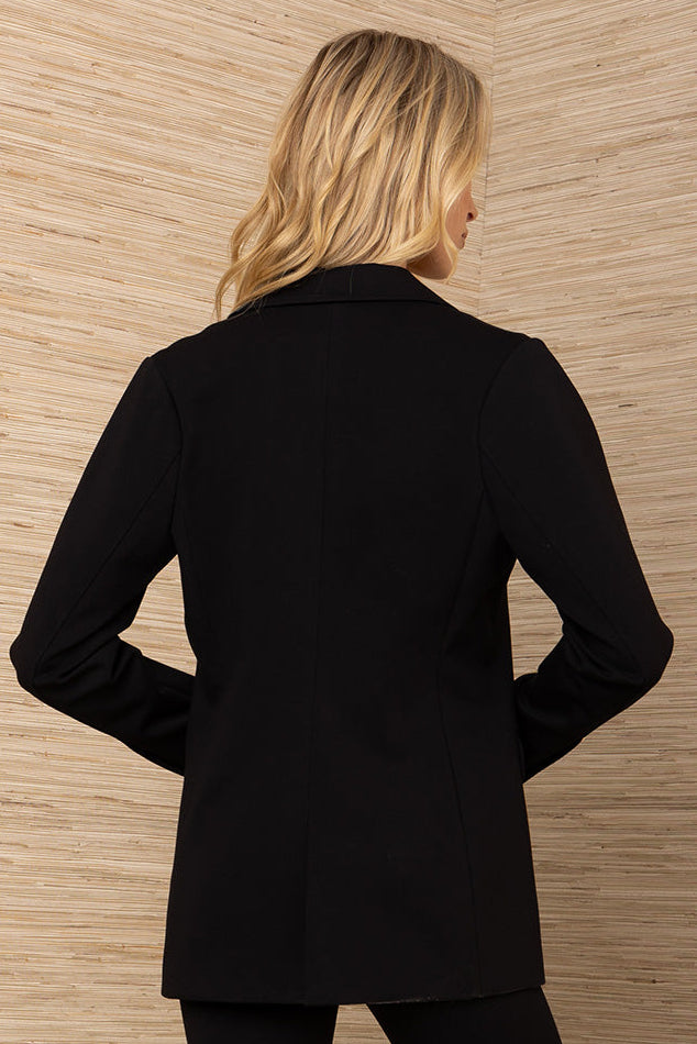 Woman wearing a black blazer standing against a wooden paneled wall.