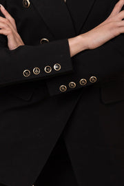 Woman wearing a black suit with gold buttons against a wooden wall.