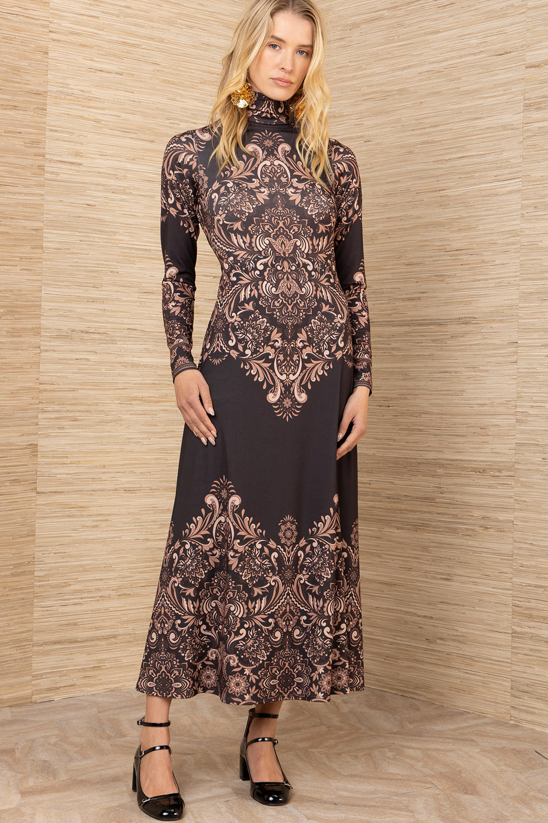 Woman wearing a long, patterned dress against a wooden paneled wall.