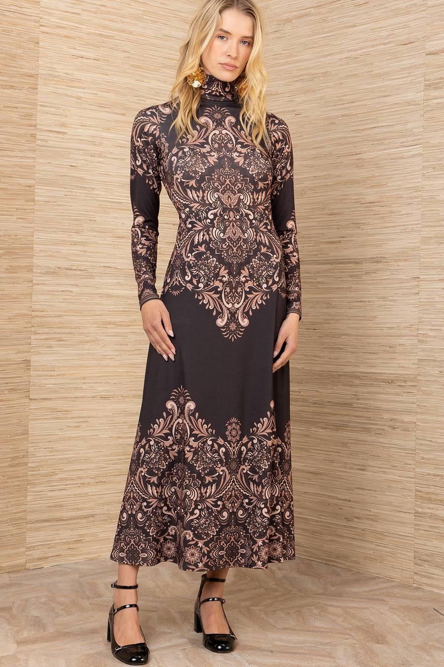 Woman wearing a long, patterned dress against a wooden paneled wall.