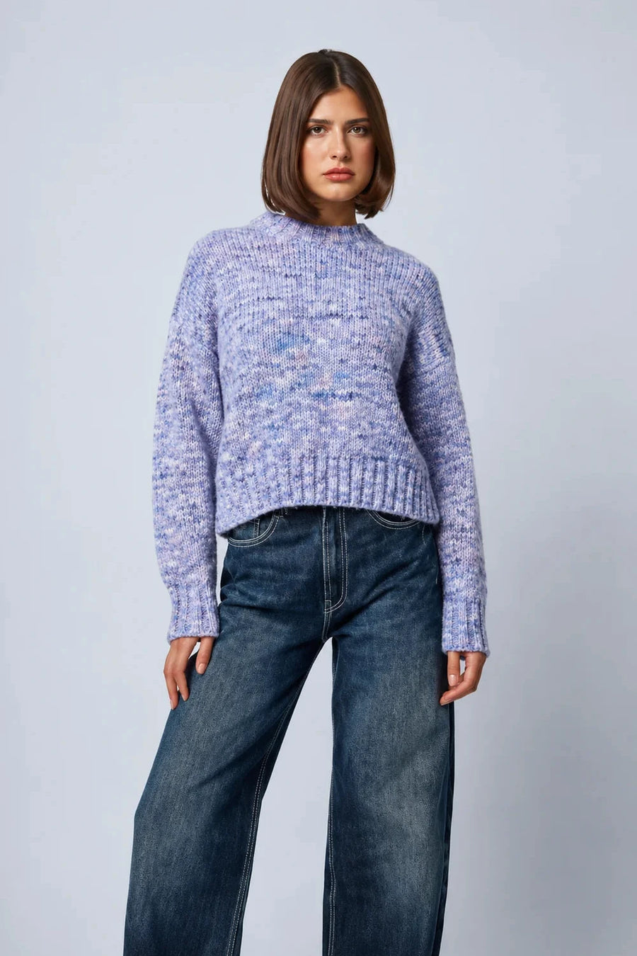 A woman modeling a light purple marled knit mock-neck sweater with dark blue wide-leg jeans, standing against a light gray studio background.