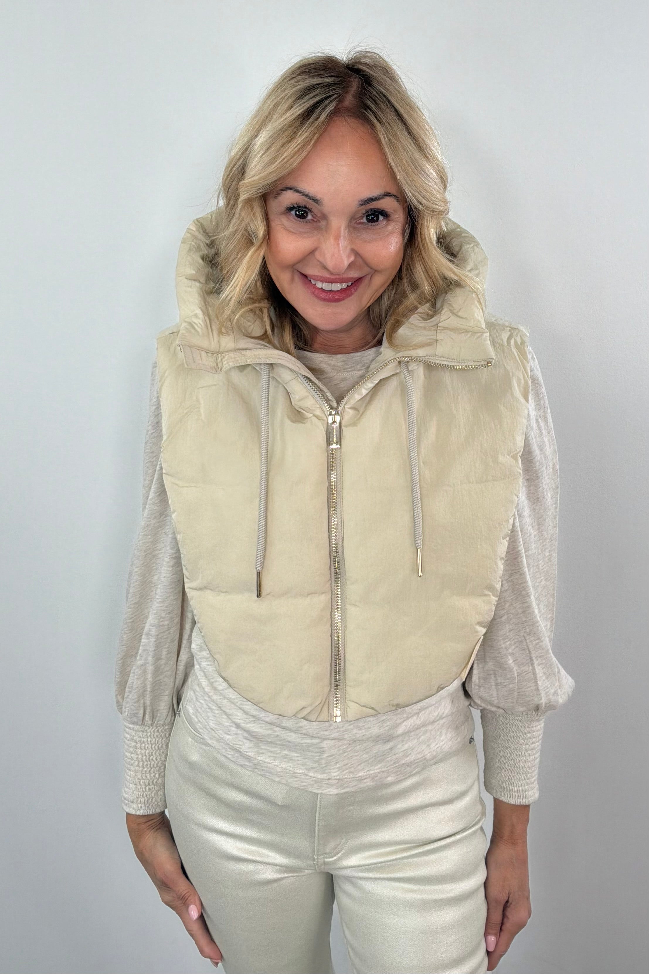 Woman wearing a beige puffer hood over a light-colored outfit against a plain background