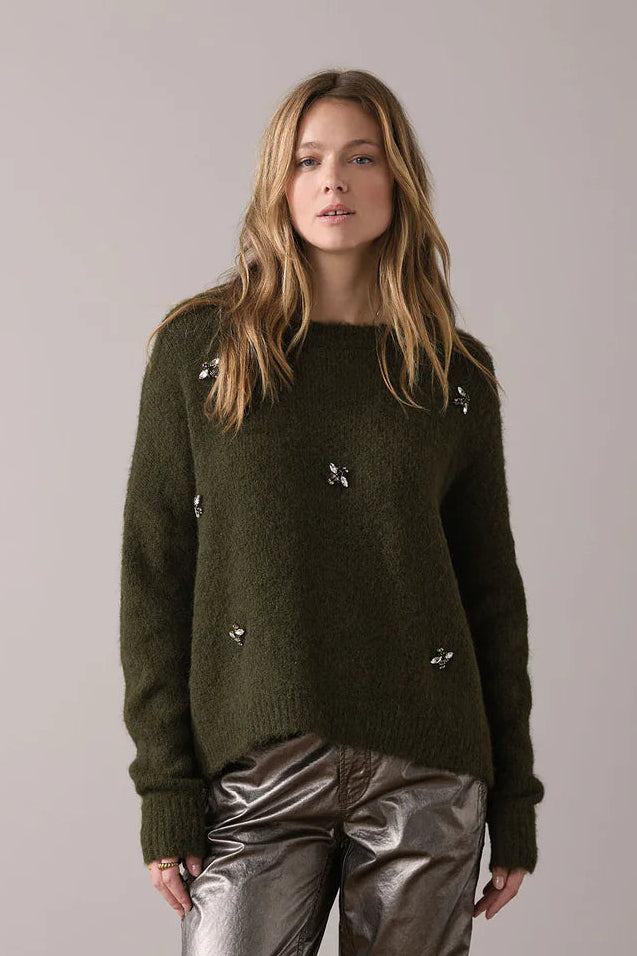 Woman wearing a dark green sweater and shiny silver pants standing on a wooden floor with a plain wall background