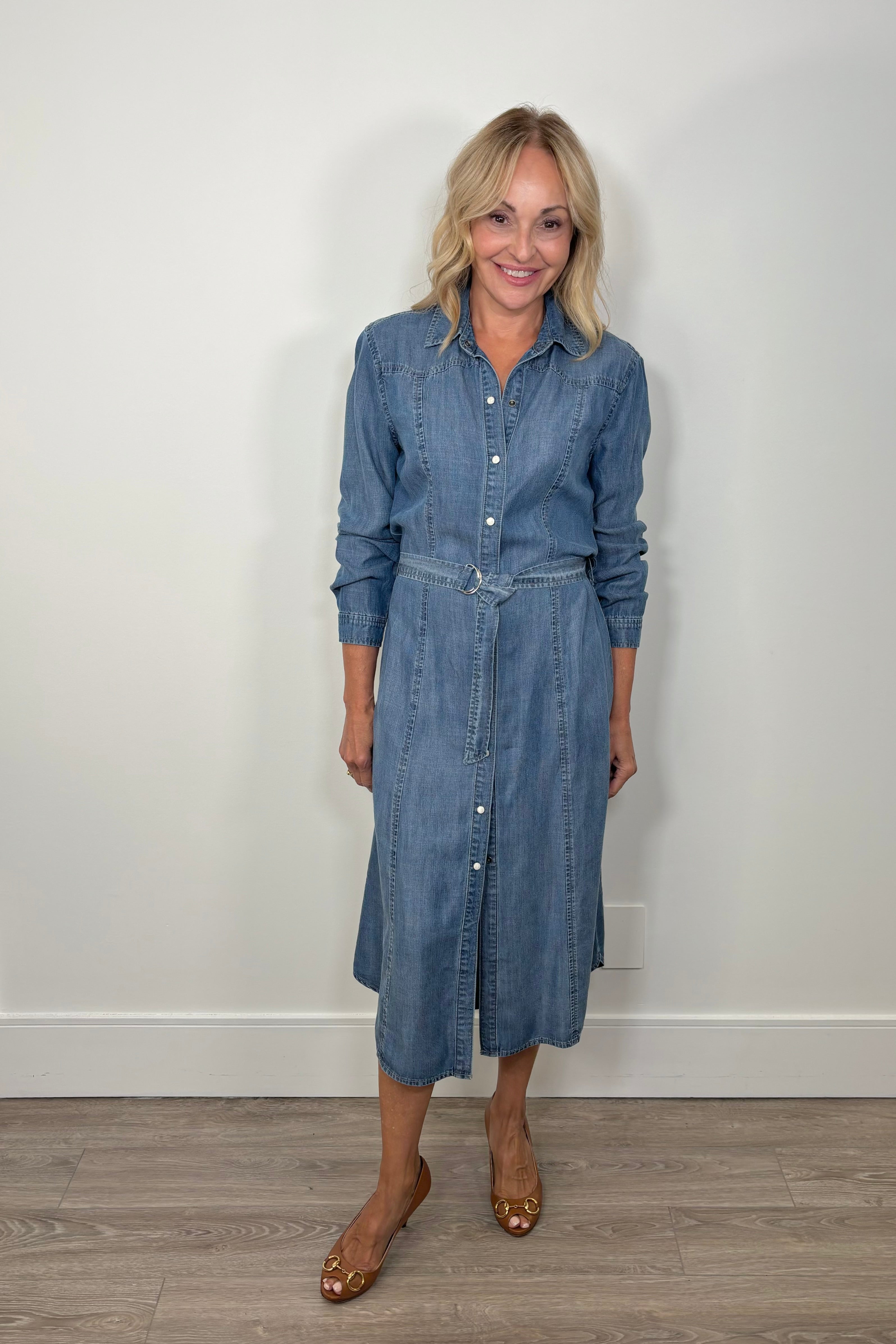 Woman wearing a blue denim belted button up, long sleeve dress against a white wall.