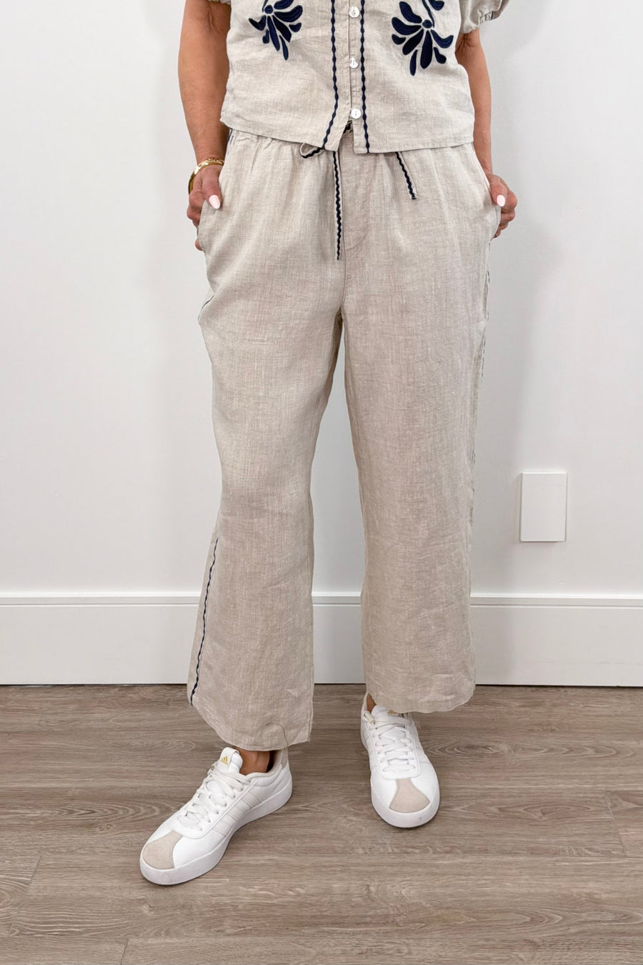 Woman wearing cream linen drawstring pants with contrast embroidery along the sides, front view