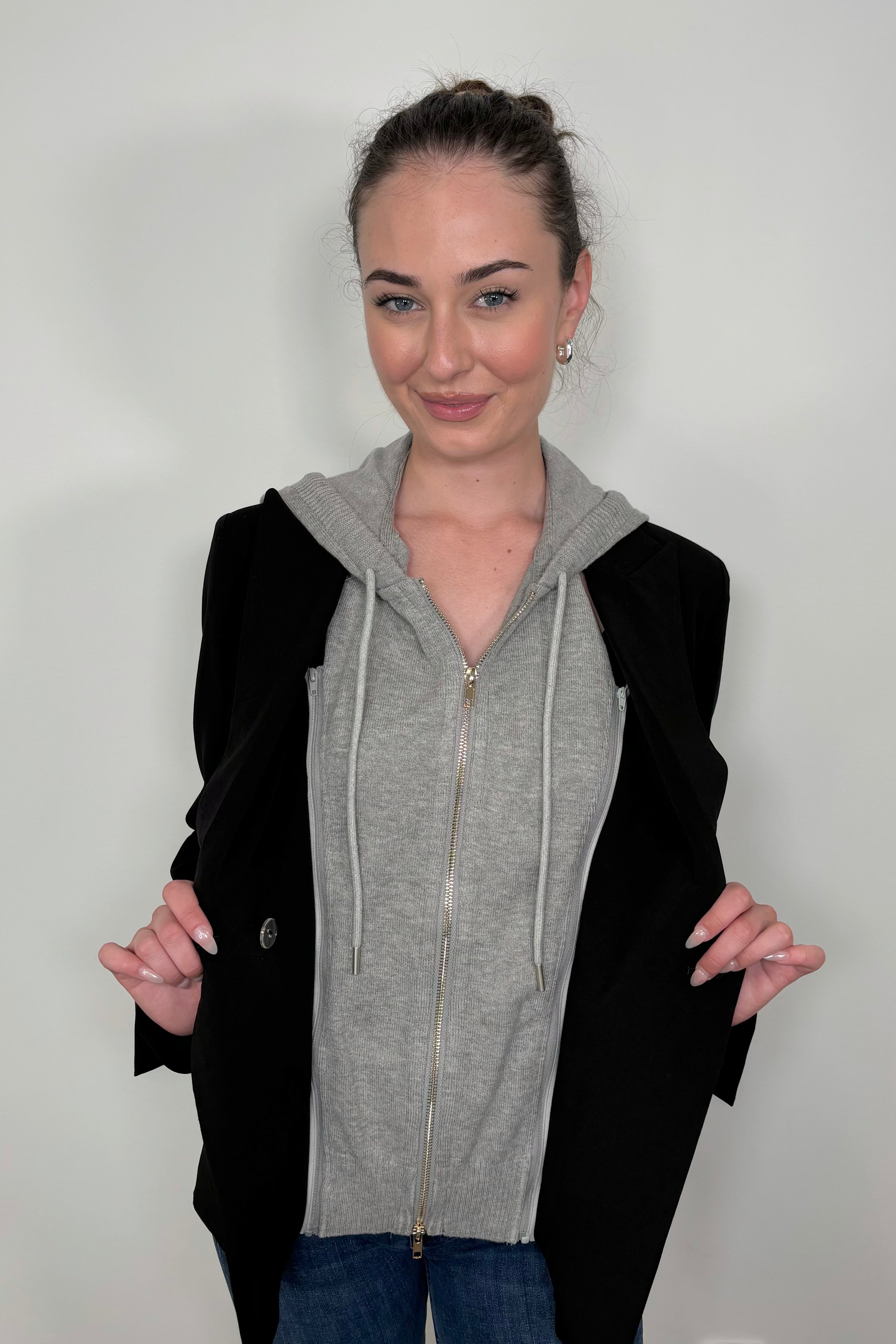 Woman wearing a black jacket that includes a detachable gray hoodie against a plain background