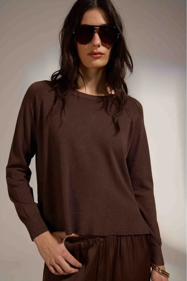 A woman wearing a dark brown long-sleeve knit top and matching brown bottoms, styled with dark sunglasses and gold bracelets, set against a neutral studio background.
