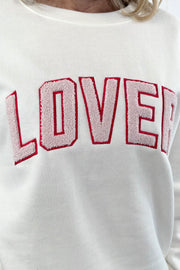 Close up of A woman wearing an off white crewneck sweatshirt with the word  “LOVER” written in pink chenille