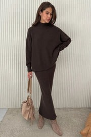 Woman wearing a dark brown outfit with a beige handbag against a light background